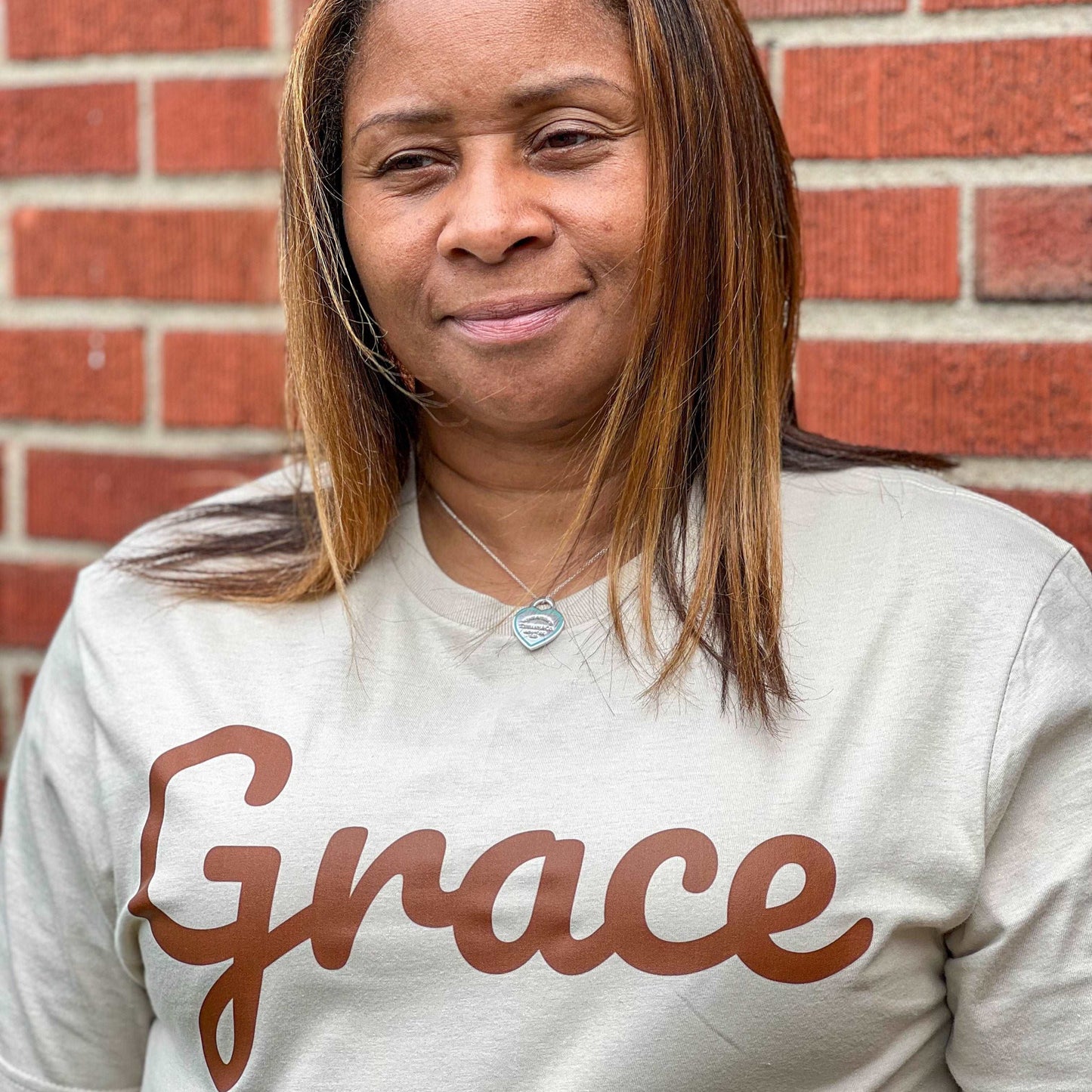 Smiling woman with shoulder-length brown hair wearing a beige t-shirt with ‘Grace’ in brown script, standing against a brick wall.