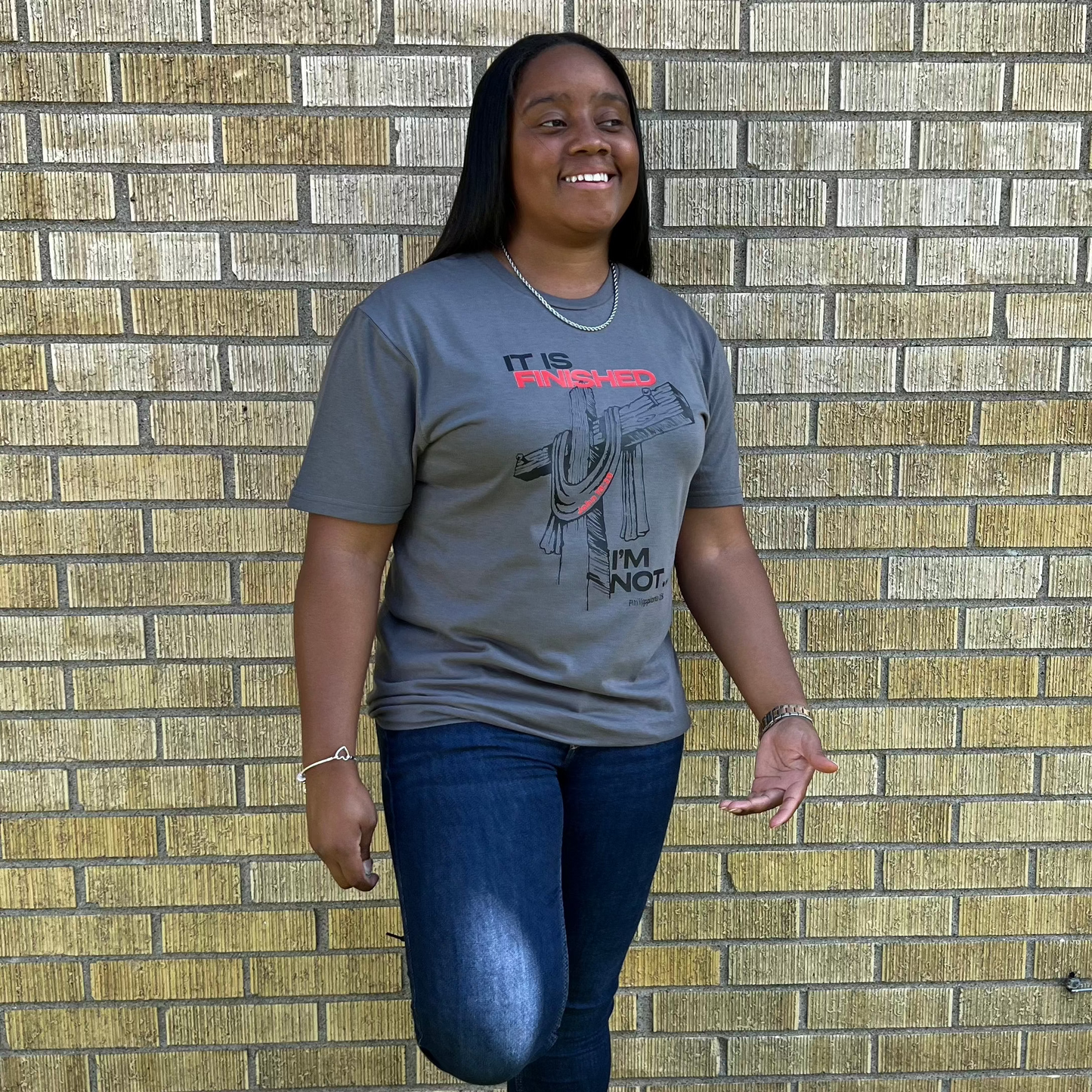 Smiling woman with long black hair wearing a gray t-shirt with a cross graphic and the words ‘It is finished, I’m not,’ standing against a yellow brick wall.