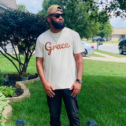 Man with a beard wearing sunglasses, a beige t-shirt with ‘Grace’ in brown script, black pants, and gold accessories, standing outdoors in a suburban setting.