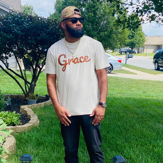 Man with a beard wearing sunglasses, a beige t-shirt with ‘Grace’ in brown script, black pants, and gold accessories, standing outdoors in a suburban setting.