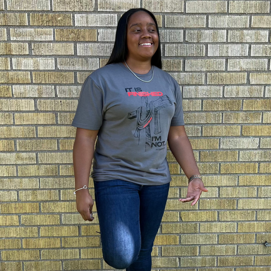 Smiling woman with long black hair wearing a gray t-shirt with a cross graphic and the words ‘It is finished, I’m not,’ standing against a yellow brick wall.